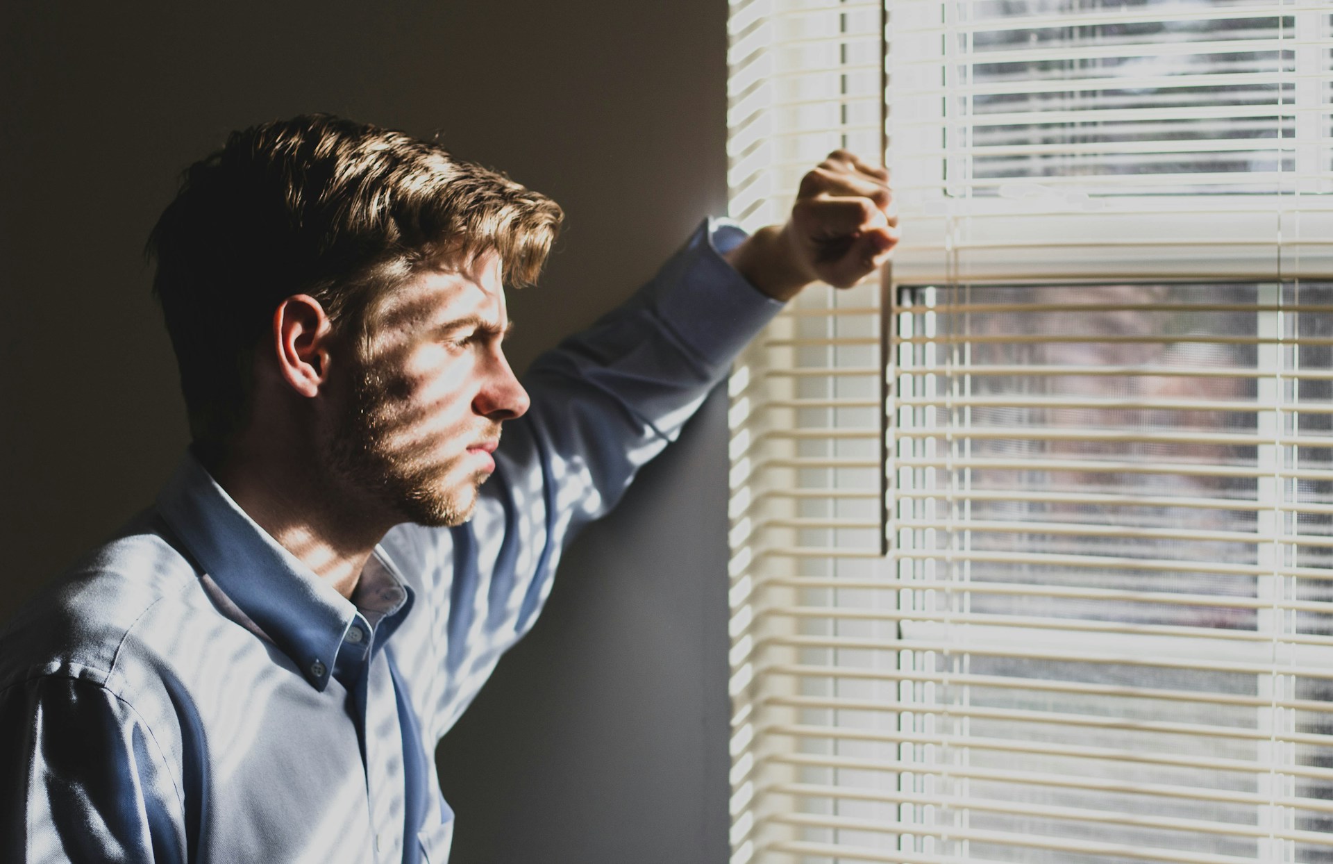 Man leaning against wall looking out the window with shades open